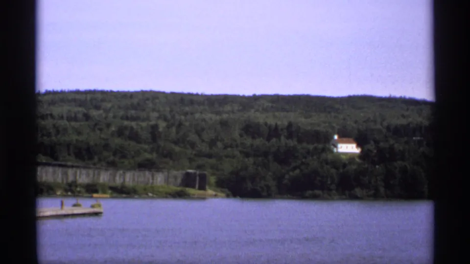 1971 Isle Royale frame showing a broad lake view, wooded hills, a partial dock, palisade sections, and a small white building on a distant slope.
