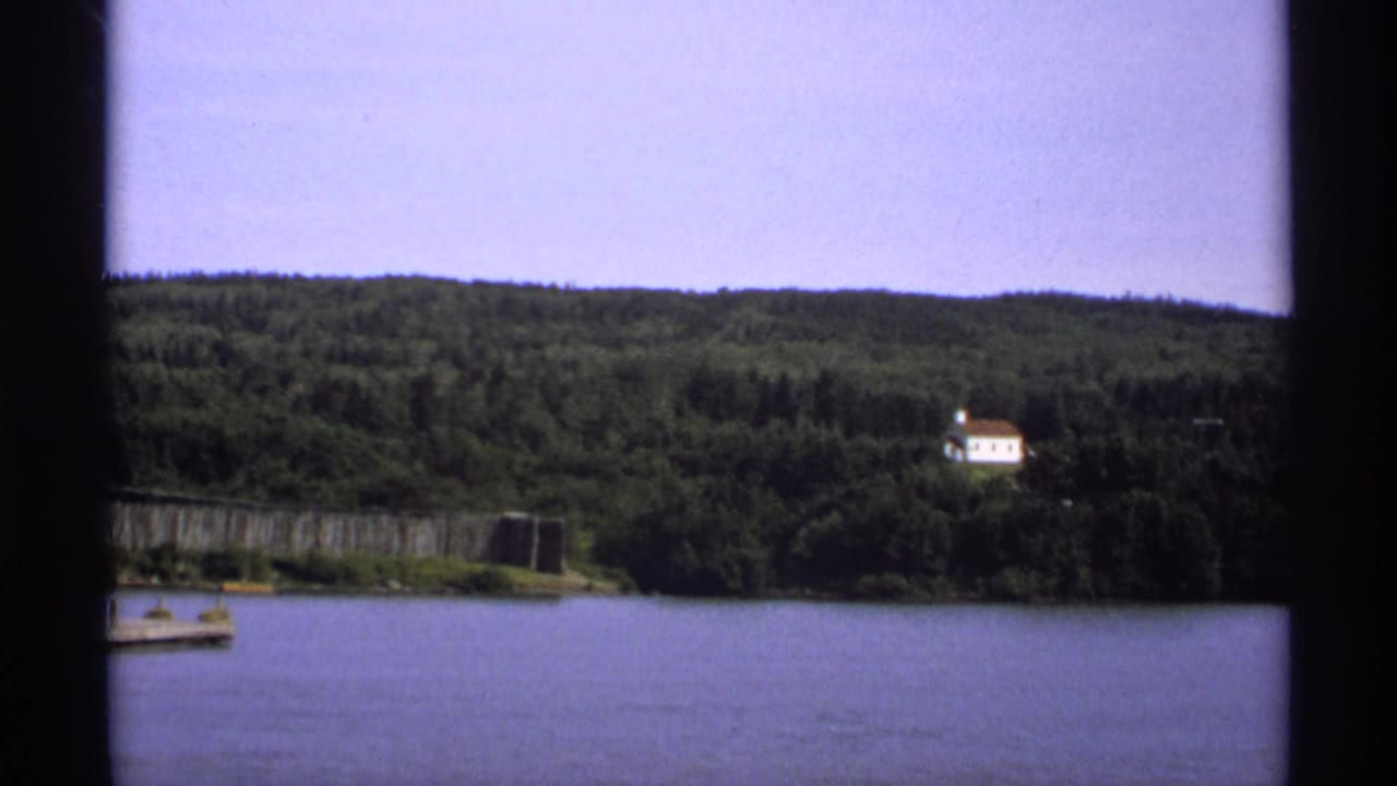 1971 Isle Royale home-movie frame showing calm lake water, forested hillside, dock edge, and a distant white house.