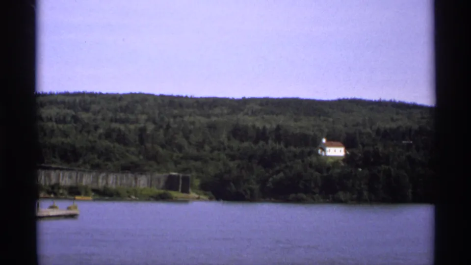 1971 Isle Royale home-movie frame showing calm lake water, forested hillside, dock edge, and a distant white house.