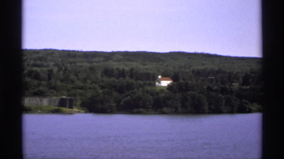 1971 Isle Royale lake view with wooded hills, blue water, and a small white house visible beyond the shoreline.