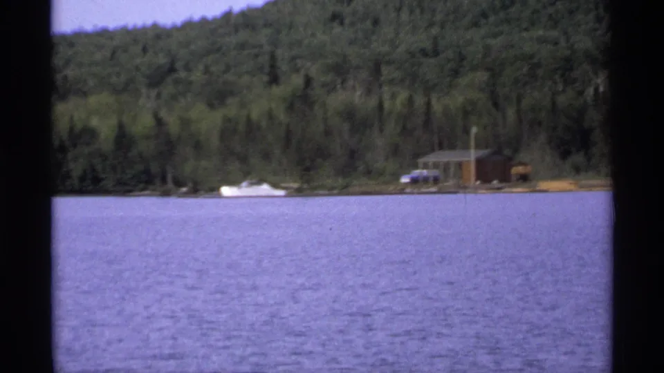 1971 Isle Royale shoreline frame showing a rustic cabin, white boat, parked car, trees, and lake water.