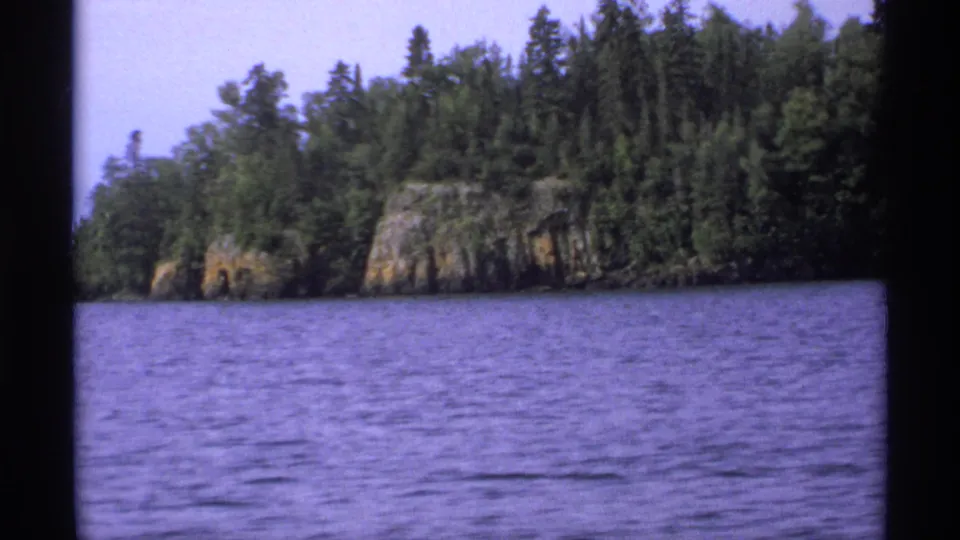 1971 Isle Royale shoreline view with rippled water, evergreen trees, and a pale rocky outcrop across the lake.