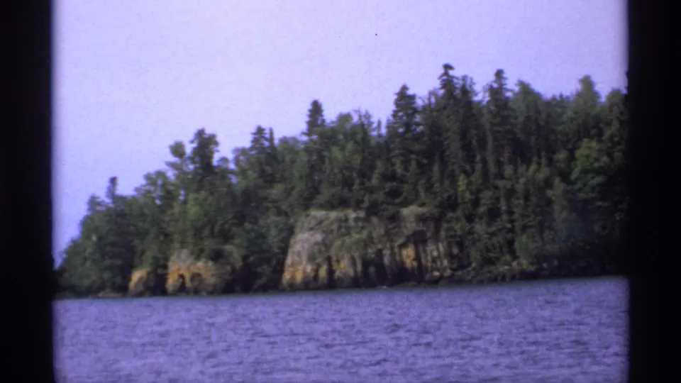 1971 Isle Royale frame of purple-tinted lake water and a forested rocky shoreline, filmed from across the water.