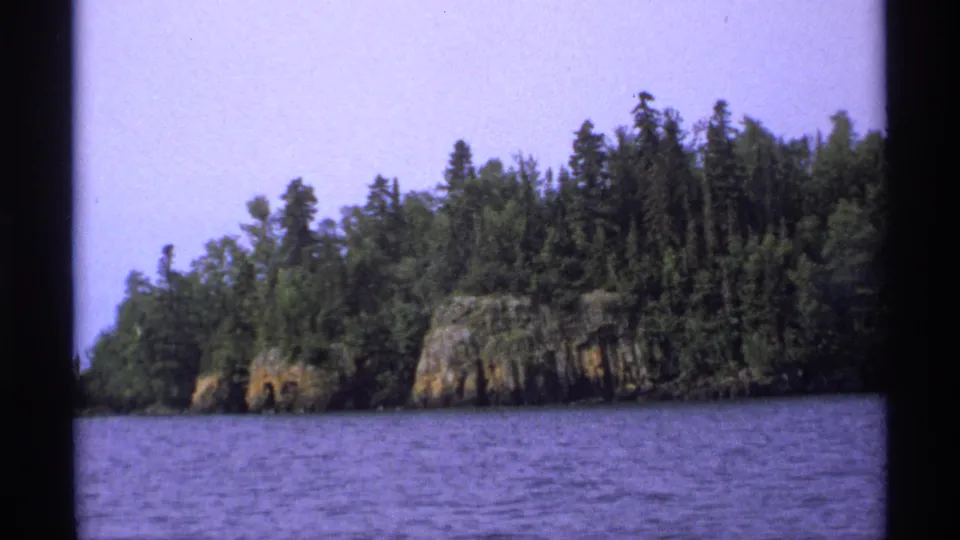 1971 Isle Royale home-movie view of a rocky, evergreen-covered shoreline across rippled lake water.