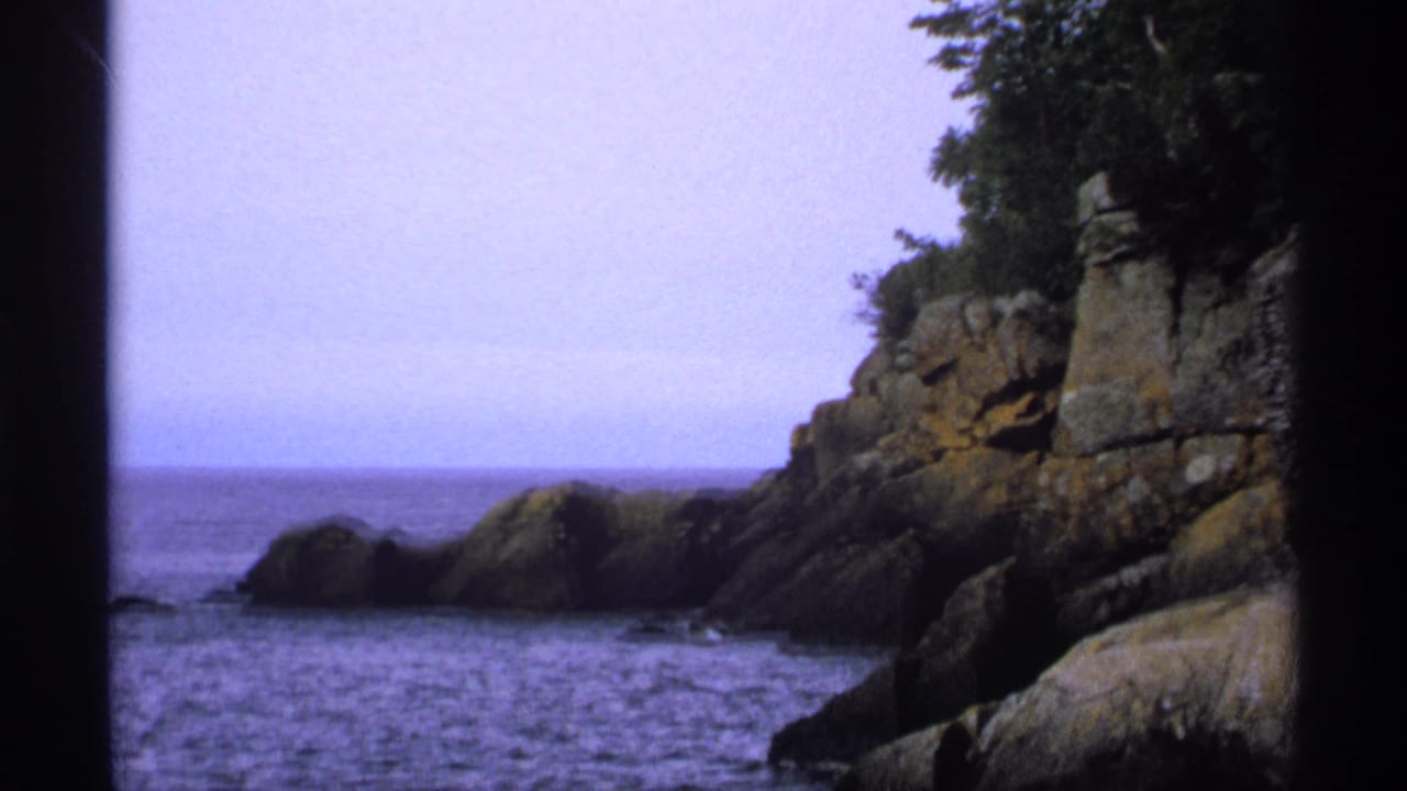 1971 8mm view of Isle Royale shoreline with calm lake water, large rock formations, evergreens, and soft blue-purple film color.