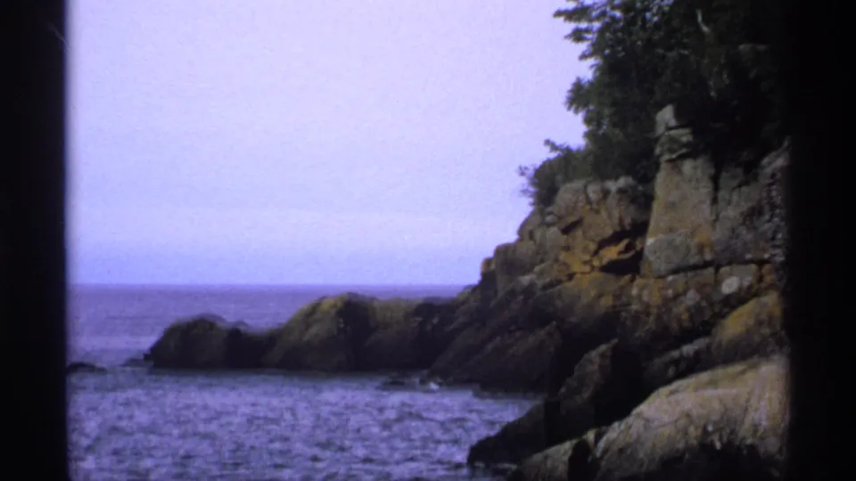 1971 8mm view of Isle Royale shoreline with calm lake water, large rock formations, evergreens, and soft blue-purple film color.