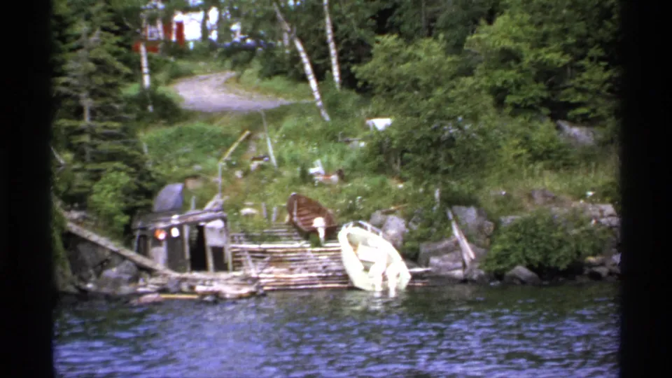 1971 Isle Royale frame showing a wooden dock, moored boats, grassy slope, trees, and distant lakeside buildings.