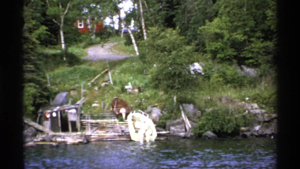 1971 Isle Royale 8mm frame of a docked white boat, rough wooden pier, green shoreline, and red buildings in the background.