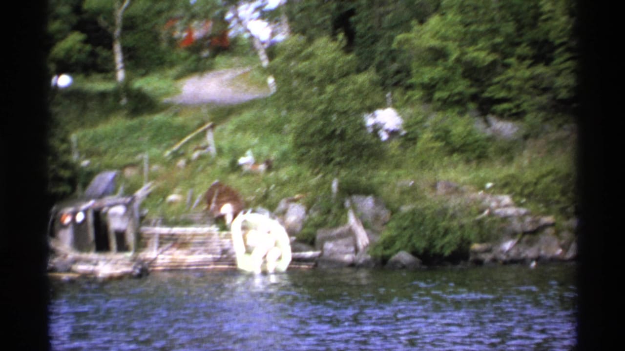 1971 Isle Royale 8mm frame showing a wooded lakeshore with a rustic dock, small boat, rocks, grassy bank, and rippling water.