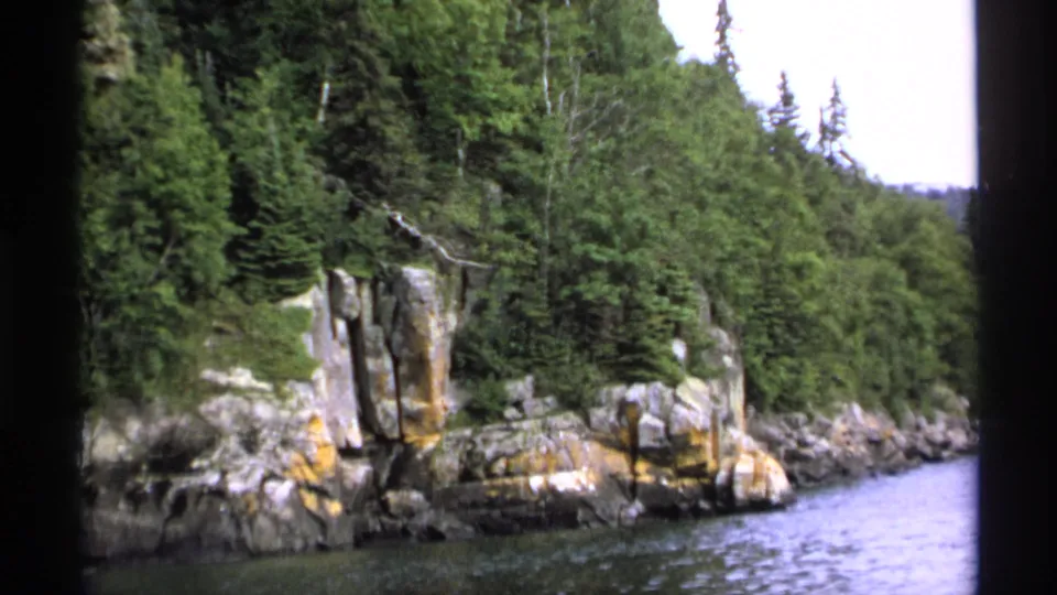 1971 Isle Royale shoreline view with layered rocks, green forest, and calm lake water filmed from a moving vantage point.
