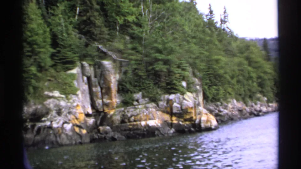 1971 Isle Royale 8mm frame of a rugged rock outcrop, forested slope, and lake water along the island shoreline.