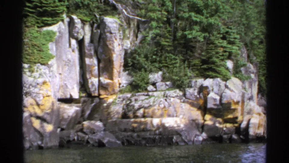 1971 boat-view frame of Isle Royale rocks and trees, with calm water below and dark vertical scan edges visible.