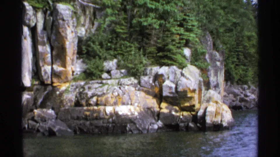 Isle Royale rocky coast viewed from water, showing forested slopes, stone ledges, lichen color, and rippled lake surface.