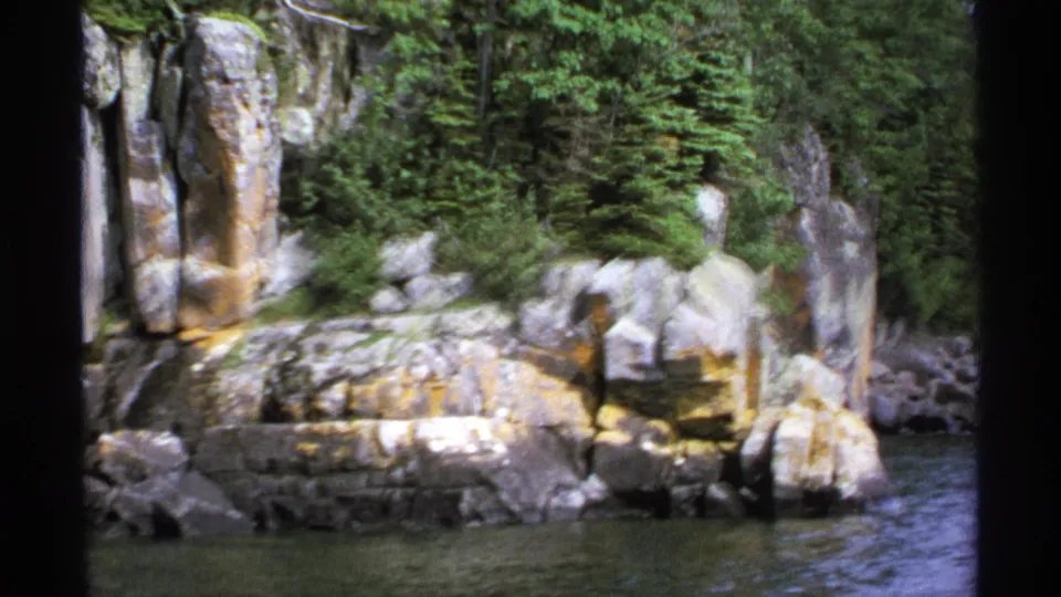 1971 Isle Royale 8mm frame showing layered shoreline rocks, yellow lichen, evergreens, and calm water from a boat-level view.