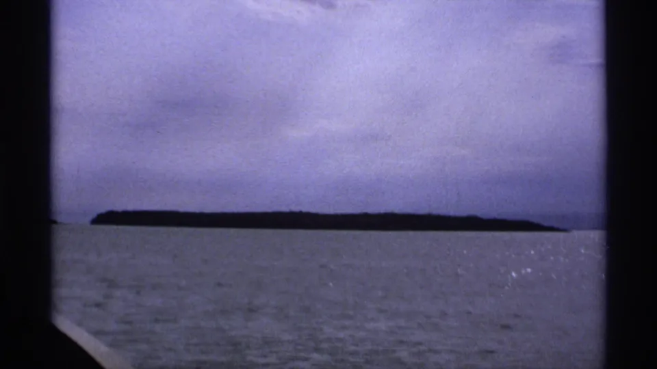 1971 Isle Royale 8mm frame with a long, dark island silhouette beyond calm lake water and a cloudy sky.