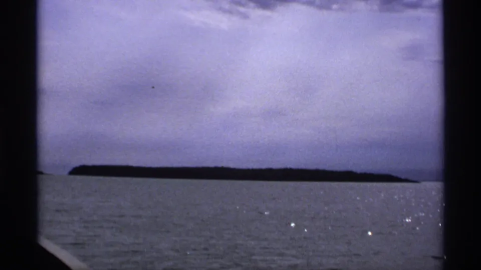 1971 Isle Royale 8mm frame showing a distant island, calm gray-blue water, small sun glints, and a cloudy sky.