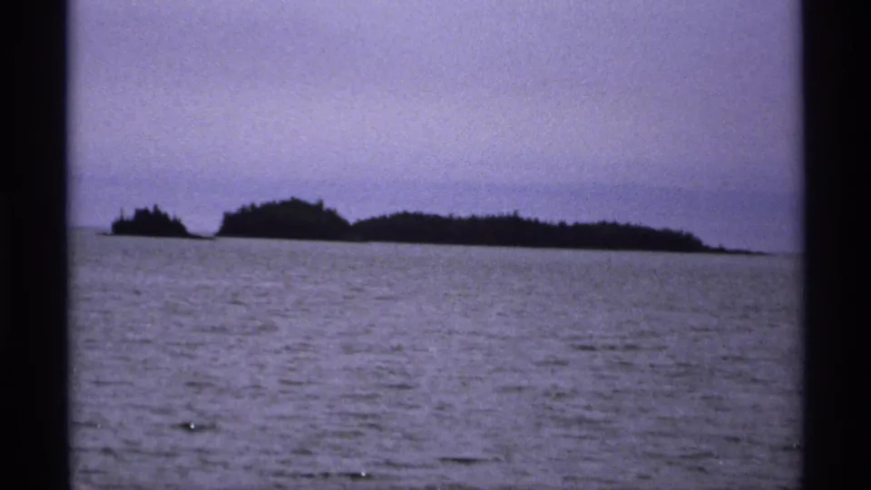1971 Isle Royale 8mm frame showing low forested island silhouettes beyond calm open water under a hazy purple-blue sky.