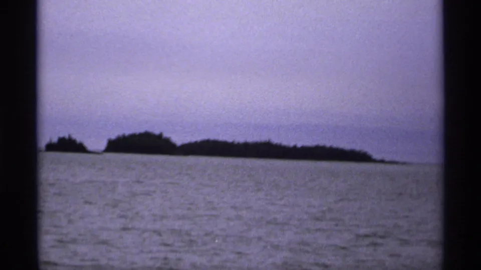 1971 Isle Royale boat-view frame with dark wooded islands stretching across the horizon over rippled lake water.