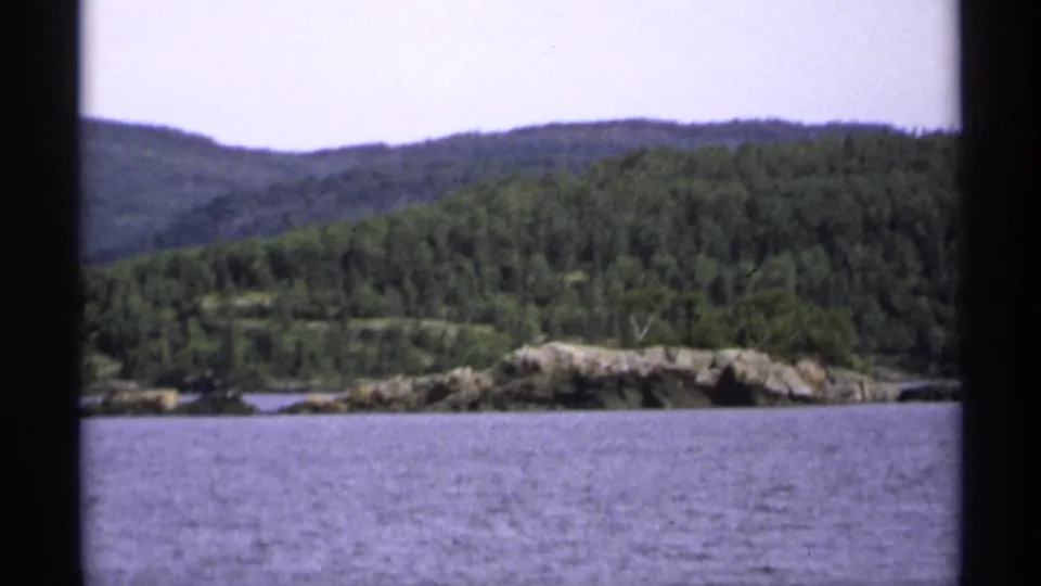 1971 Isle Royale frame showing a closer rocky shoreline with dense green forest and rolling hills beyond the water.