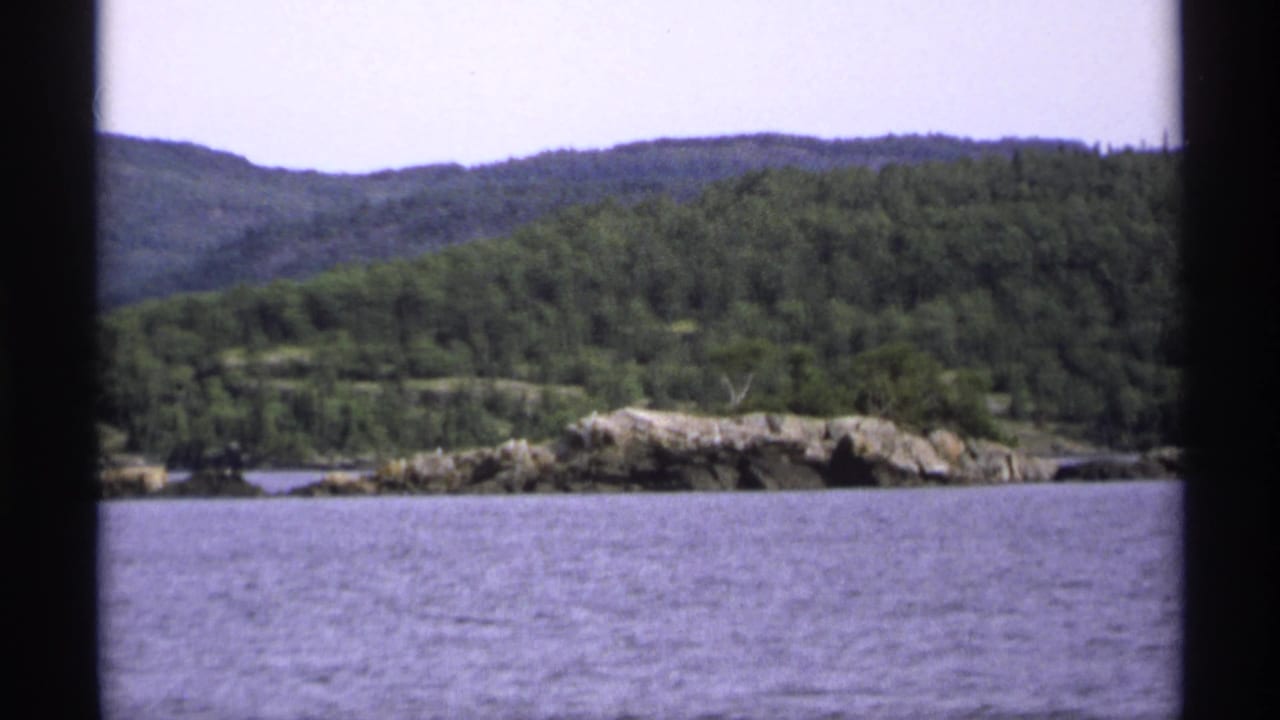 1971 Isle Royale home-movie view of a wooded shoreline and pale rocks across calm water under an overcast sky.