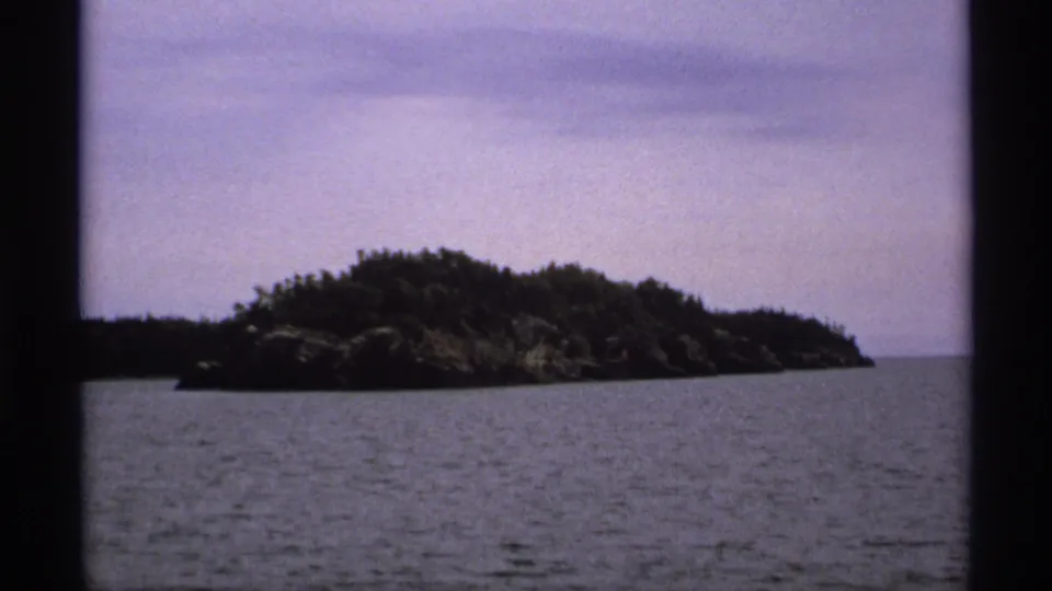 1971 Isle Royale footage showing a low wooded island and rocky shoreline across rippled Great Lakes water.