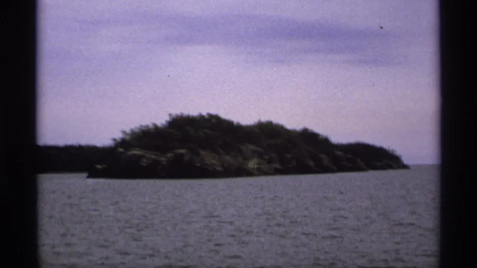 Tree-covered Isle Royale island filmed from across the water, with a rocky edge and hazy overcast sky.