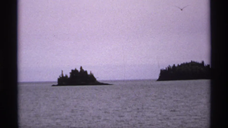 1971 Isle Royale home-movie frame showing wooded islands silhouetted across calm water beneath a muted purple-blue sky.