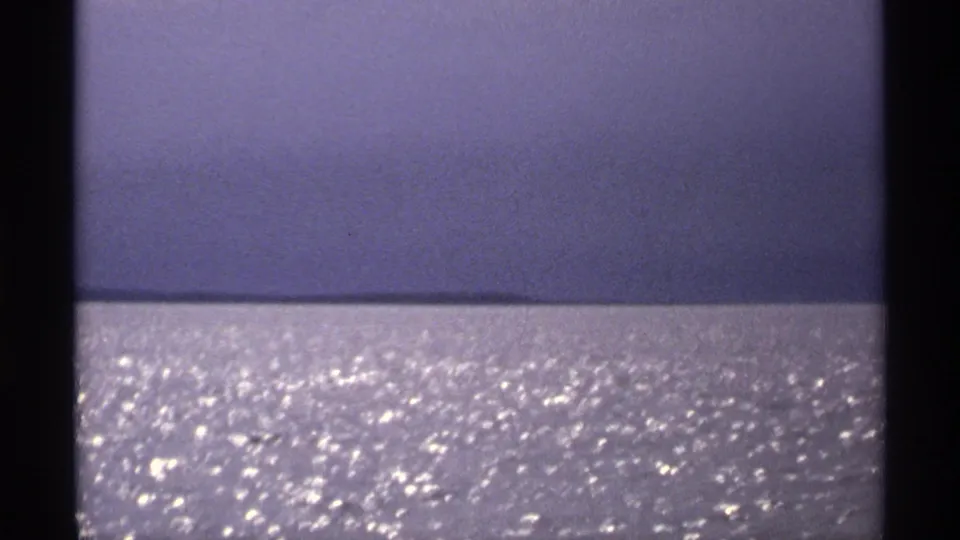 1971 Isle Royale frame showing open water with strong sun glitter and a hazy distant shoreline or horizon under blue-purple sky.