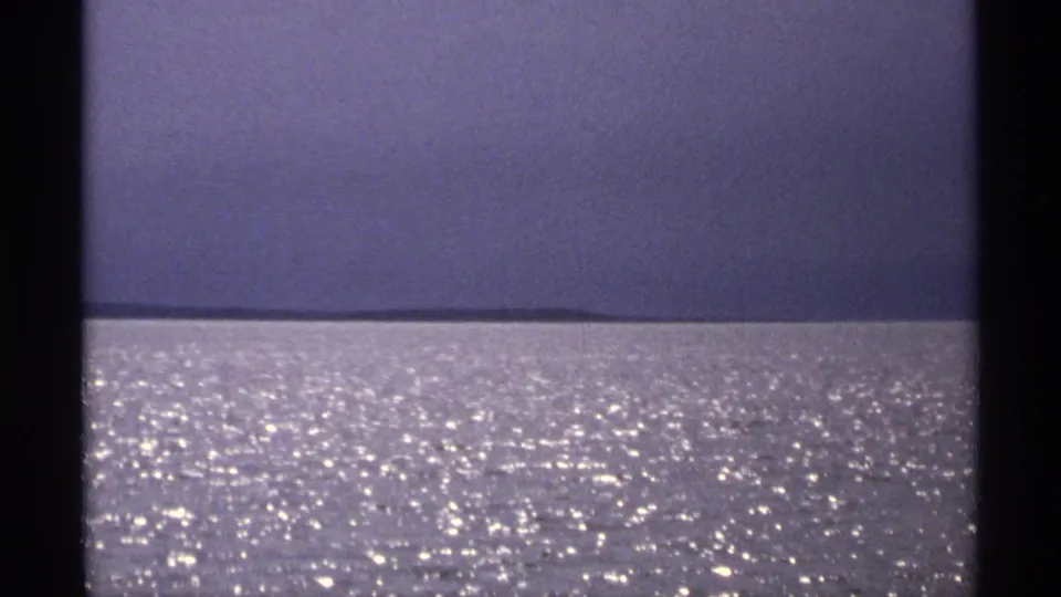 1971 Isle Royale home-movie frame showing rippling open water, bright surface reflections, and a far low landmass under a pale sky.