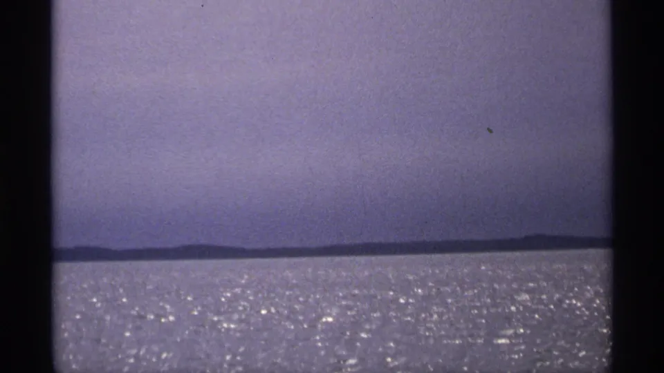 1971 Isle Royale frame with a low horizon line, faint shoreline, pale sky, and bright water texture from 8mm home-movie footage.