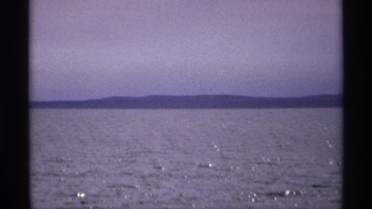 1971 8mm view of calm water near Isle Royale, Michigan, with low land across the horizon and soft purple-blue sky.