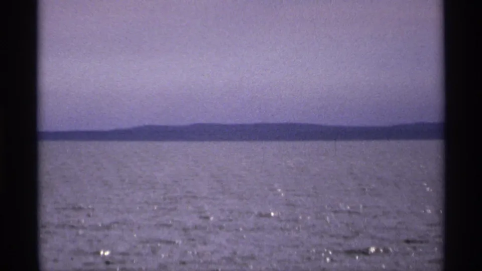 1971 8mm view of calm water near Isle Royale, Michigan, with low land across the horizon and soft purple-blue sky.