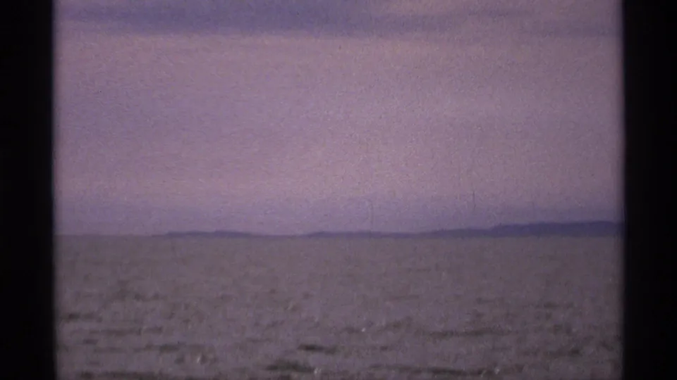 Final 8mm frame of calm Isle Royale water with a dark, low shoreline and pale purple-blue sky.