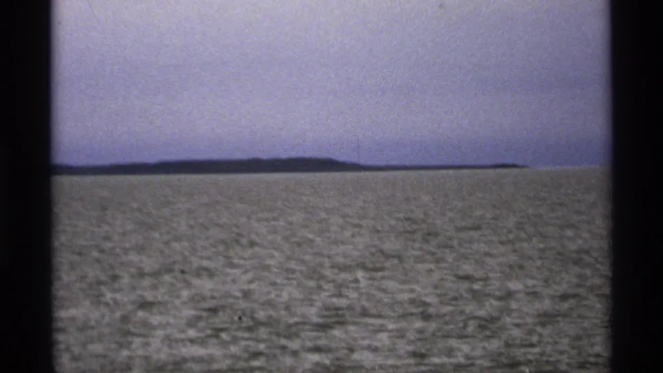1971 8mm Isle Royale-area shoreline seen across rippled lake water, with faded color and visible black film borders.