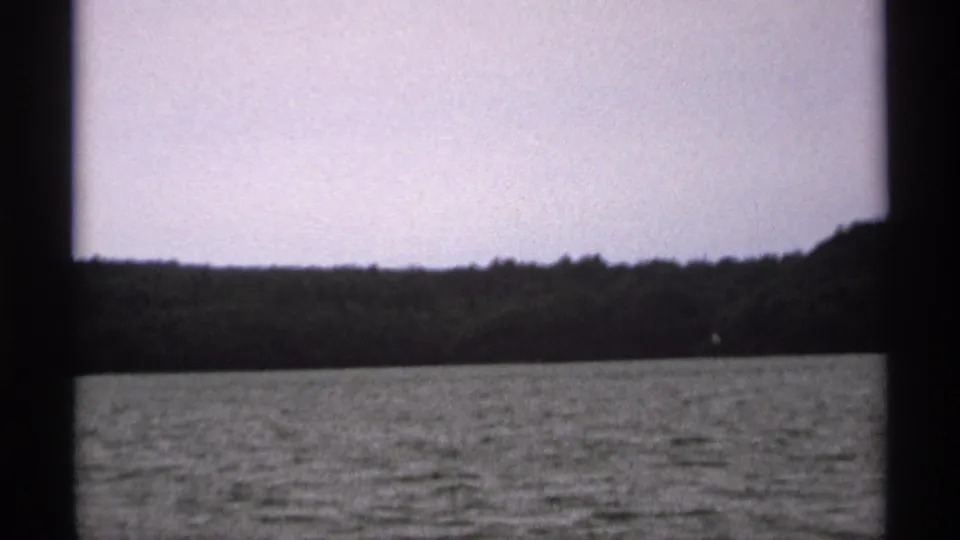 1971 home-movie frame of Isle Royale from the water, with a distant wooded shore and muted sky visible through the film gate.