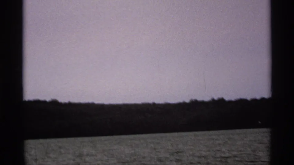 1971 8mm boat-view frame near Isle Royale, with the wooded shoreline tilted slightly above rippled lake water.