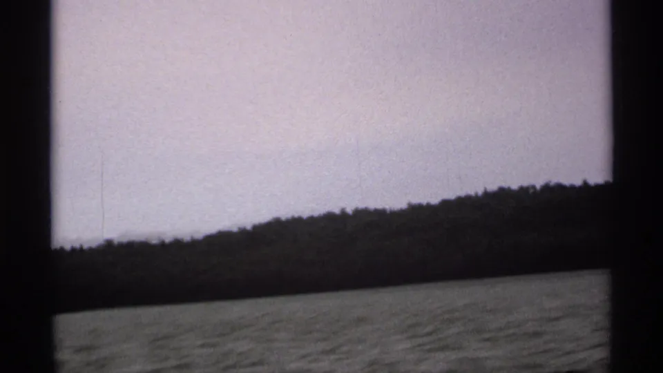1971 Isle Royale 8mm frame showing a low wooded slope beyond lake water, filmed from a moving boat.