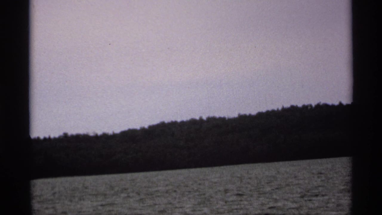 1971 Isle Royale home-movie frame showing rippled lake water, a wooded shore, pale sky, and dark vertical framing from a boat view.