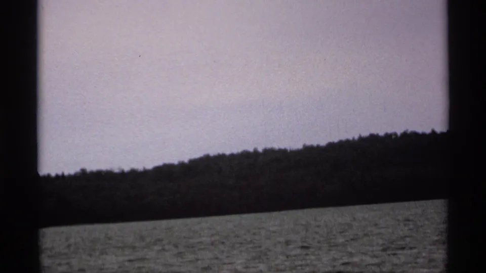 1971 Isle Royale home-movie frame showing rippled lake water, a wooded shore, pale sky, and dark vertical framing from a boat view.