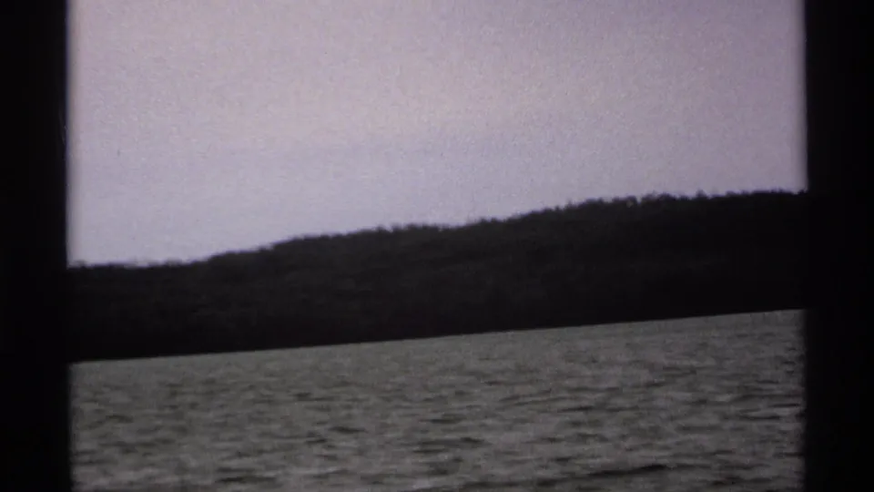 1971 Isle Royale boat-view frame with calm lake water, a tree-lined shore, pale sky, and dark window-like framing in the foreground.