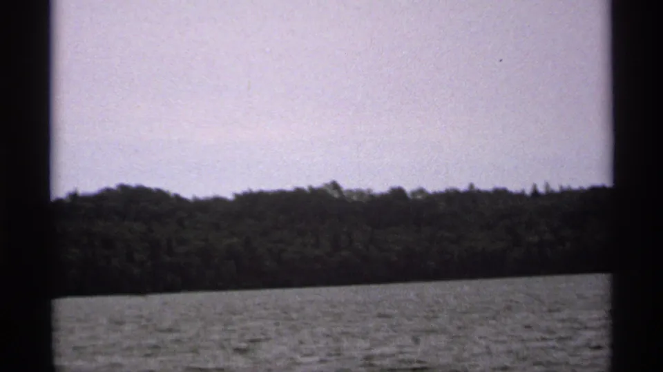 1971 Isle Royale 8mm frame showing a low forested shoreline and rippled lake water, with dark boat framing at the sides.
