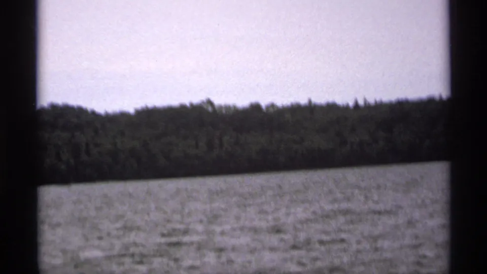 1971 Isle Royale home-movie frame with bright lake water, dense shoreline trees, pale sky, and dark boat or window framing.