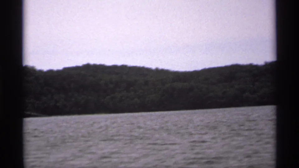 1971 Isle Royale home-movie frame showing rippled lake water, low forested hills, and a pale sky from a boat viewpoint.