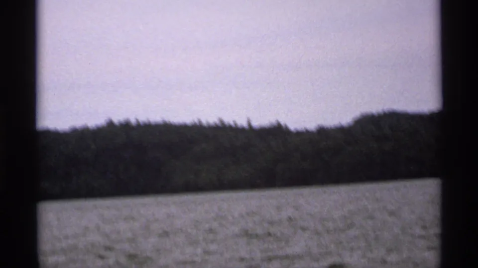 1971 Isle Royale home-movie view of rippled lake water and a tree-covered shoreline framed by dark boat-window or porthole edges.