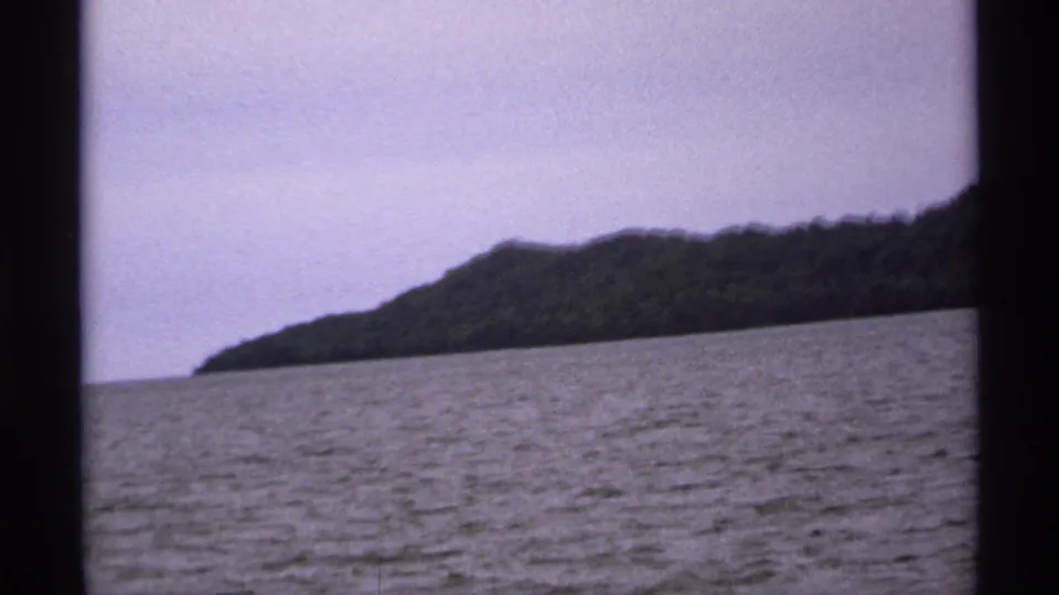 1971 Isle Royale shoreline appears across deep rippled water, with dark window framing and soft purple-gray 8mm color.