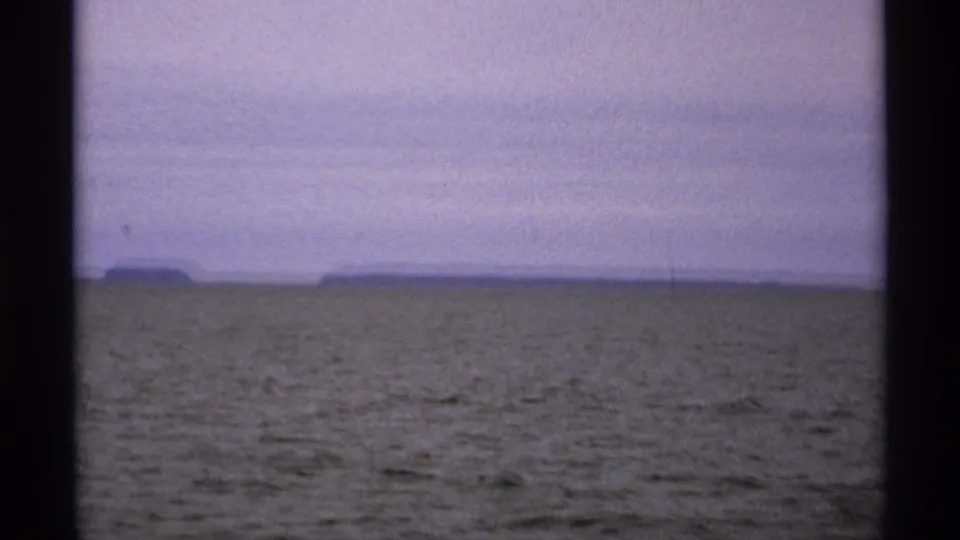 1971 Isle Royale view across calm water toward a distant shoreline, framed by dark vertical edges from the filming position.