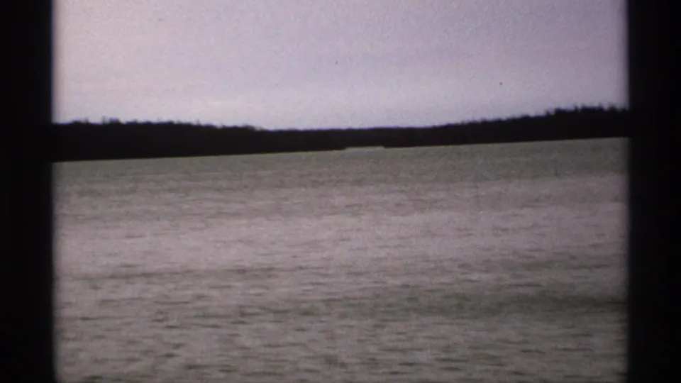 1971 Isle Royale footage shows calm water and a darker tree-lined shore, partly bordered by the camera-side frame.