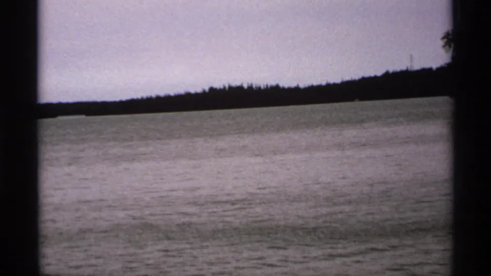 1971 Isle Royale home-movie frame showing lake water, pale sky, and a dark distant treeline viewed from a boat-like position.