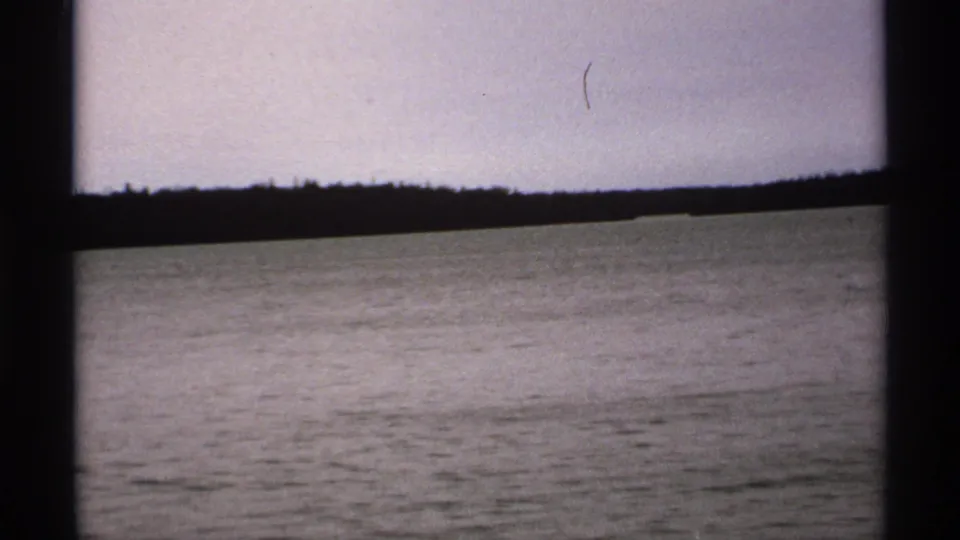 1971 Isle Royale footage frame with open water, low wooded horizon, pale sky, and dark framing at the sides.