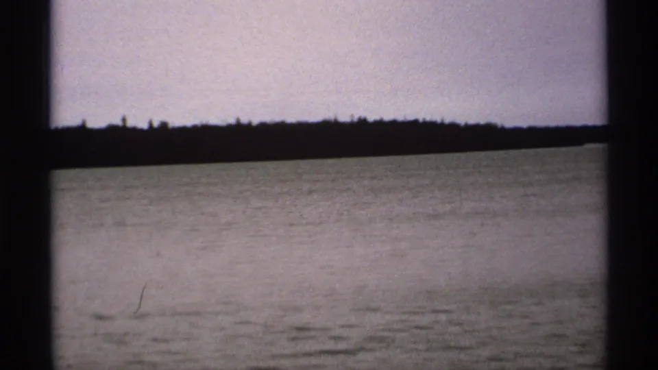 Soft 8mm frame from Isle Royale in 1971 showing rippled water, a distant tree-covered shore, and muted overcast light.